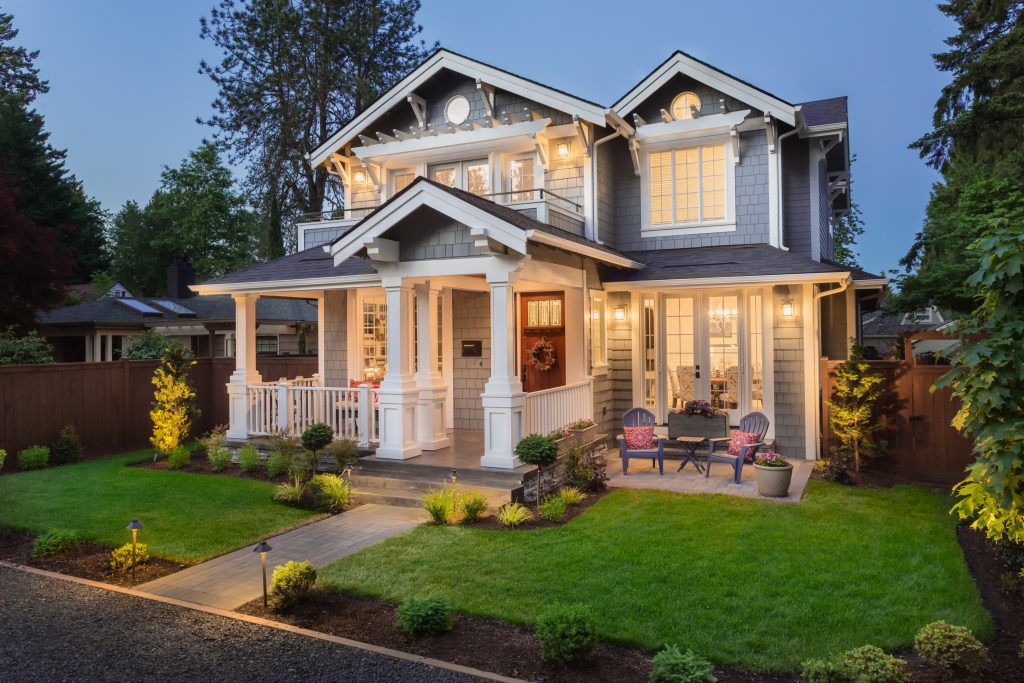 Grey and white home, lit up, in small, manicured yard with a blue sky behind it.